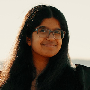 Vaishi Sistla. She is smiling and wearing a black dress and glasses. She is standing in front of a body of water and dry field.